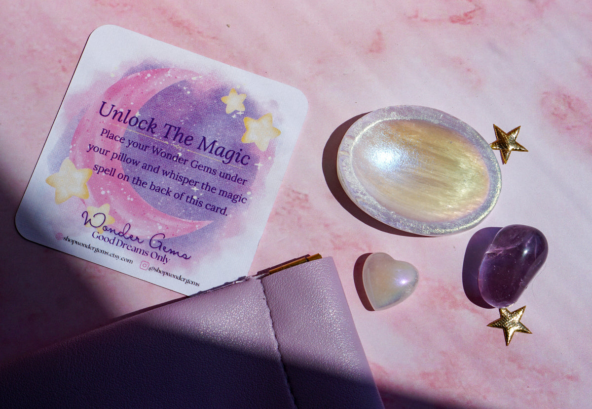A square card reading 'Unlock The Magic' with a pink and purple watercolor design and gold stars, arranged on a pink marble-like surface with a lavender pouch, a gold star, a white pearl, and a purple amethyst crystal.