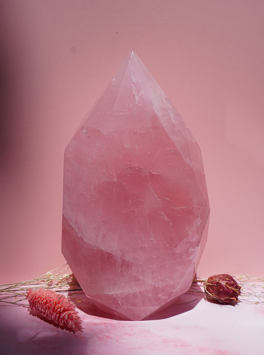 A large faceted pink crystal  faceted freeform flame sits centered on a pink surface against a pink background, with dried floral stems placed around its base.