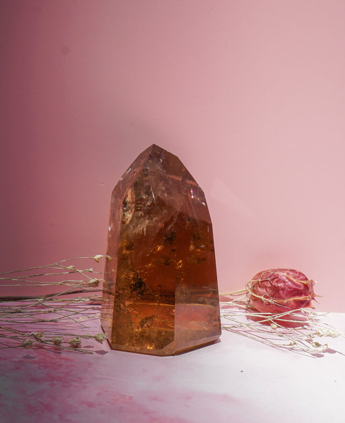 A tall, faceted honey smokey citrineamber crystal tower with internal inclusions, styled on a white surface with dried flowers against a solid pink background.