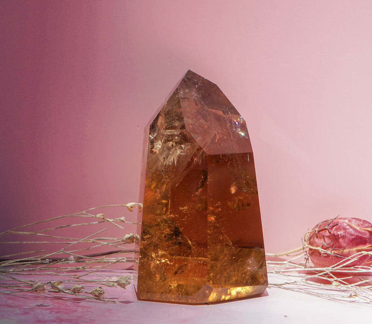 A large pointed honey smokey citrine quartz crystal tower in warm amber-orange tones with visible internal inclusions, styled on a pink surface against a pink background with dried branches and a dried flower nearby.