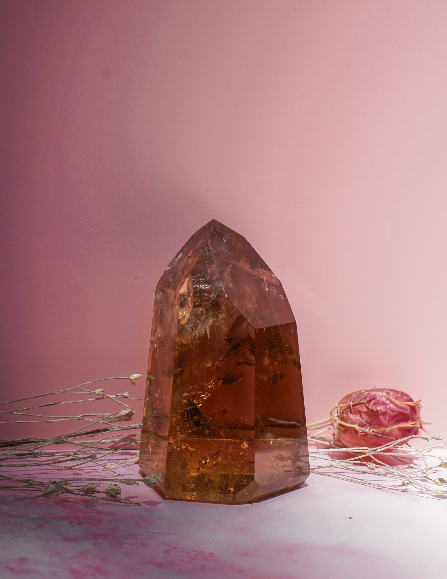 A large faceted honey smokey citrine crystal tower with dried botanical elements inside, set against a soft pink background with a dried pink rose prop.