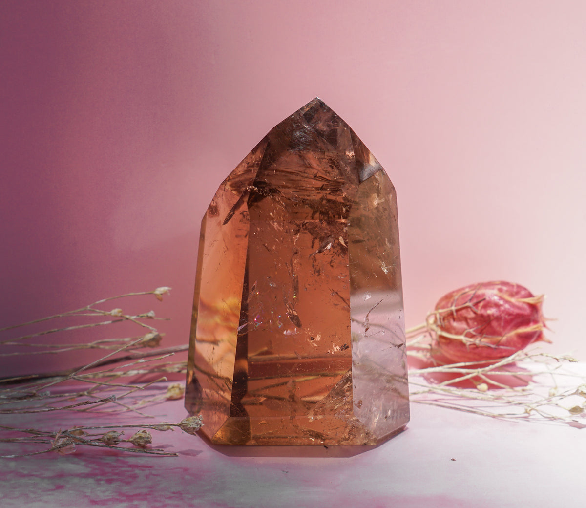 A large, polished smoky honey citrine quartz crystal point with warm amber-brown tones, styled on a pink surface with dried flowers against a soft pink background.