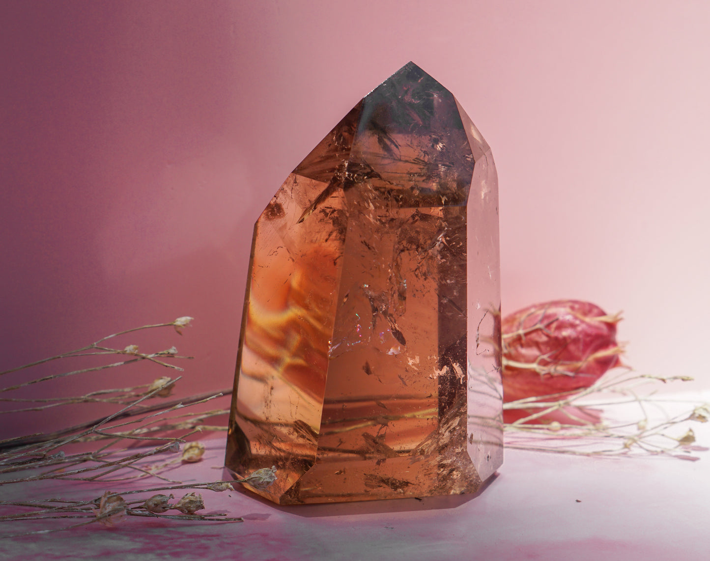 A large natural smoky honey citrine quartz crystal point with internal orange and brown banding, displayed on a pink surface with dried botanicals against a soft pink background.