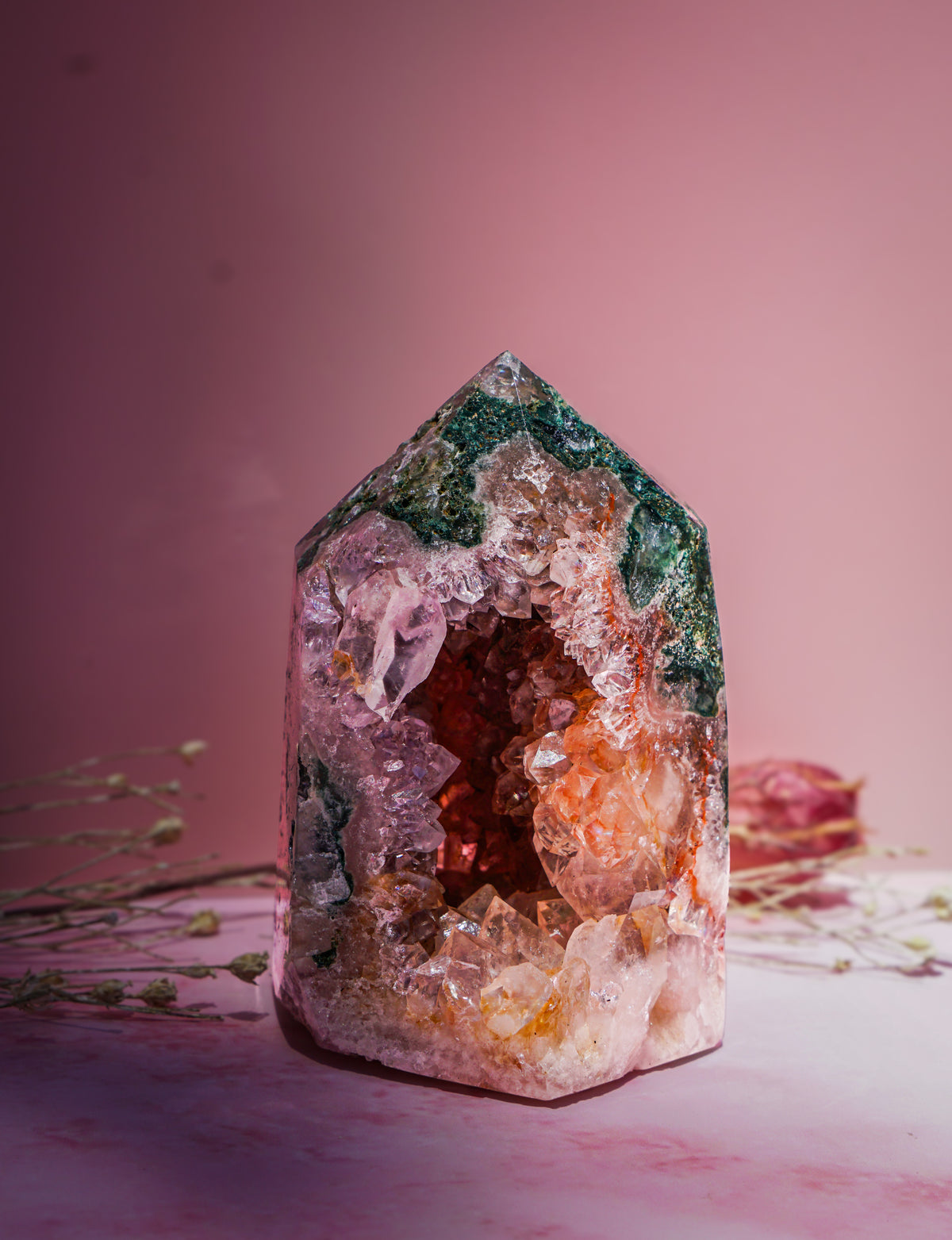 A natural  pink amethyst and jasper crystal tower with a pointed top, featuring layers of green, pink, orange, and clear quartz, displayed on a pink surface against a pink background.