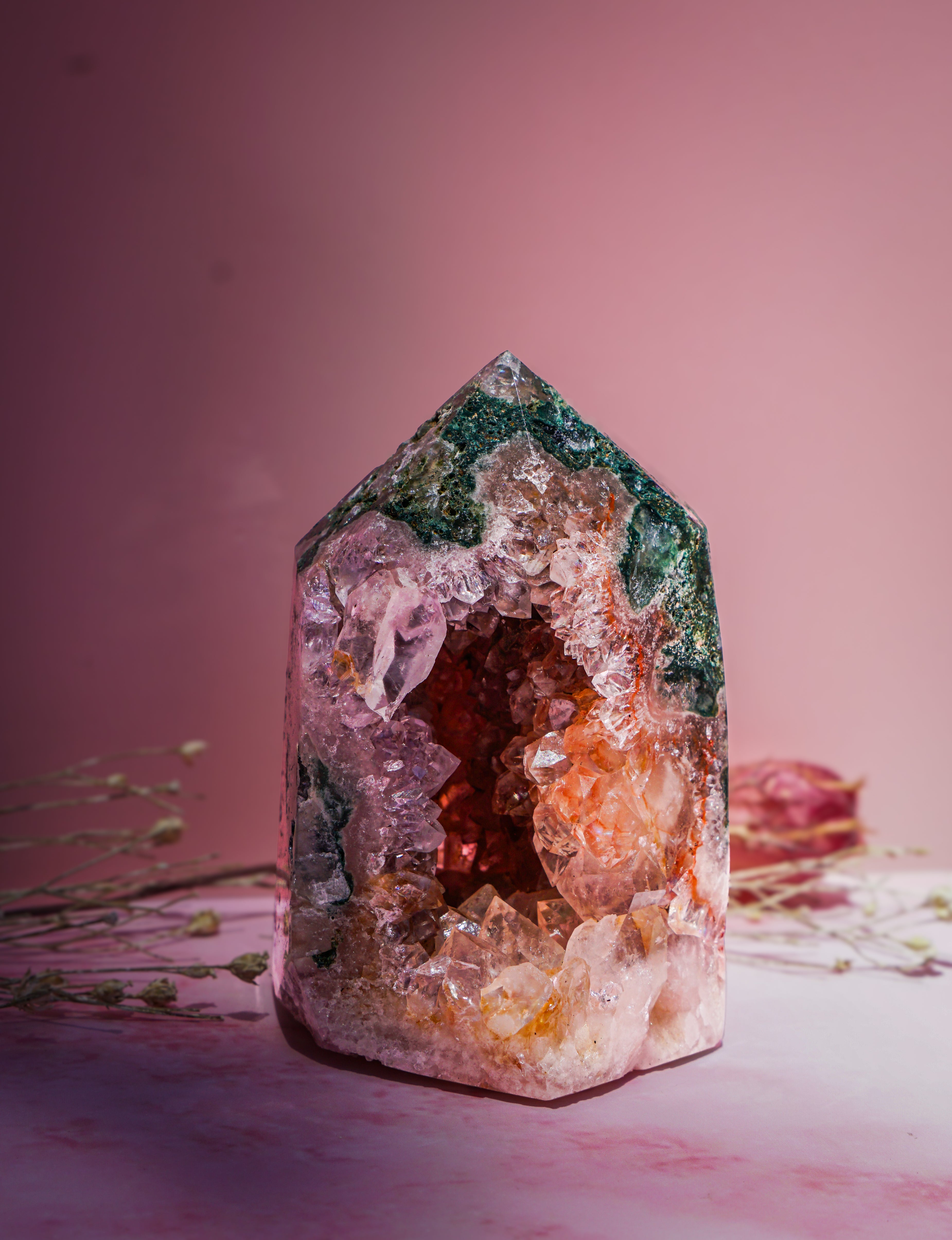 A natural  pink amethyst and jasper crystal tower with a pointed top, featuring layers of green, pink, orange, and clear quartz, displayed on a pink surface against a pink background.