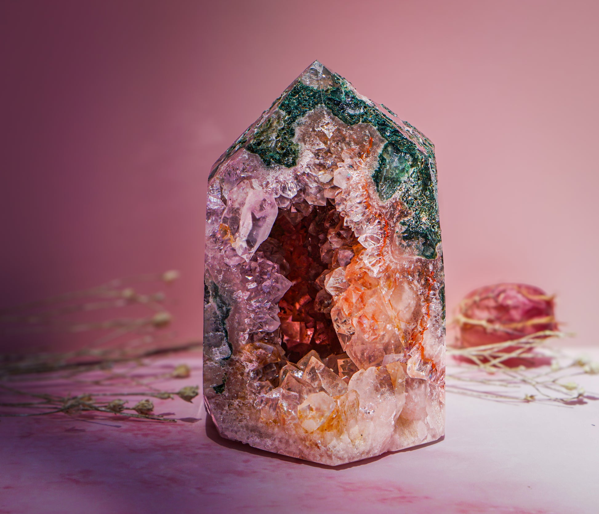 A natural pink amethyst and jasper crystal tower geode specimen with a pointed top, featuring layered clusters of clear quartz, orange-brown crystals, and green matrix, displayed on a pink surface against a pink background.
