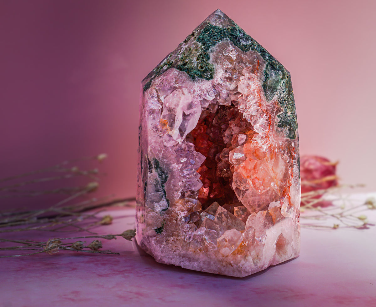 A natural  pink amethyst and jasper crystal tower with a pointed hexagonal shape, featuring a pink and white outer crystal cluster, a warm orange interior, and green matrix at the top, displayed on a pink surface with dried flowers.