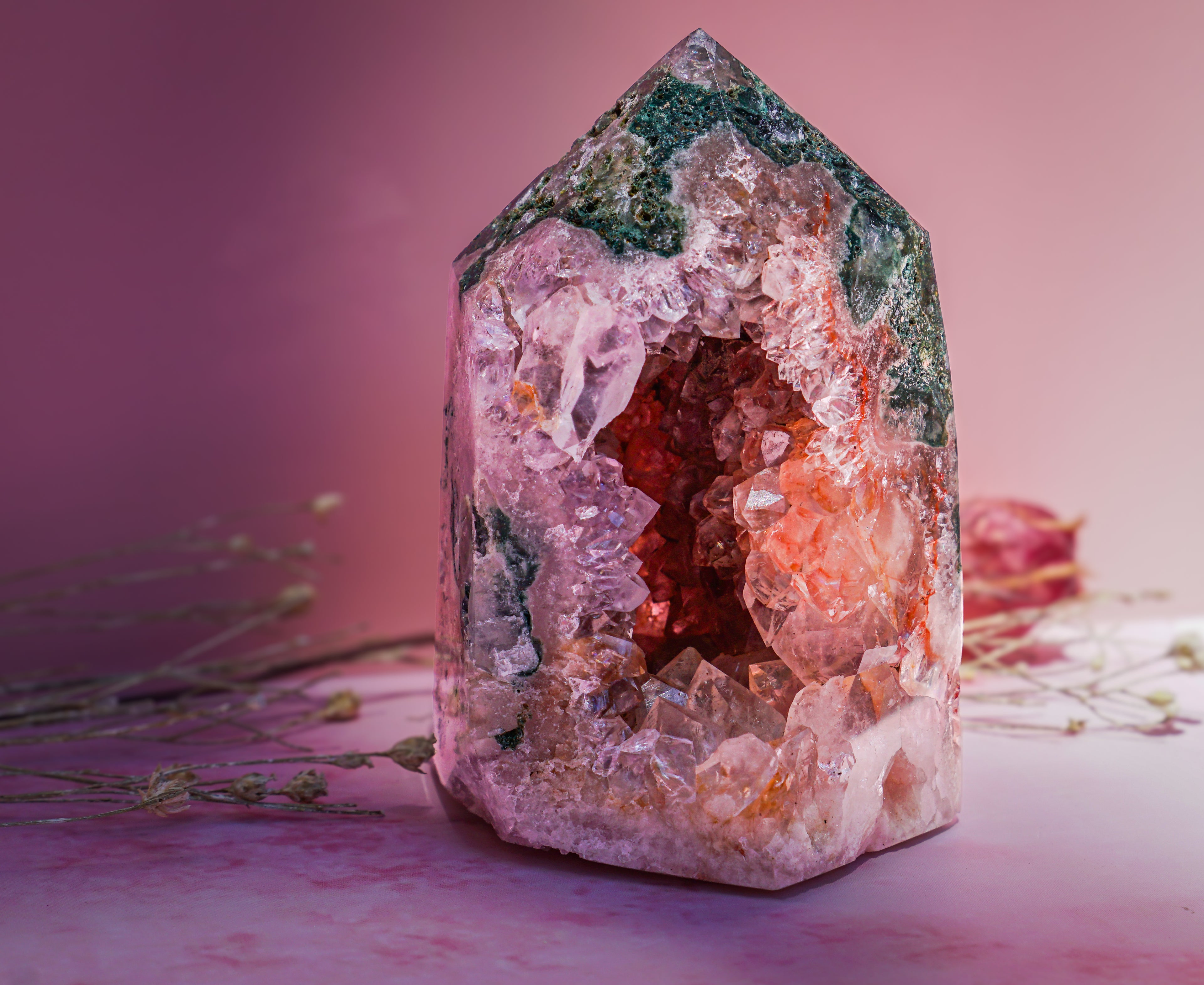 A natural  pink amethyst and jasper crystal tower with a pointed hexagonal shape, featuring a pink and white outer crystal cluster, a warm orange interior, and green matrix at the top, displayed on a pink surface with dried flowers.