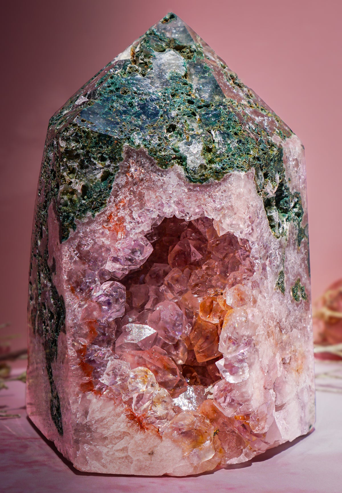 A natural pink amethyst and jasper crystal tower specimen with a pink quartz cavity, green and blue matrix, and orange inclusions, photographed against a soft pink background.
