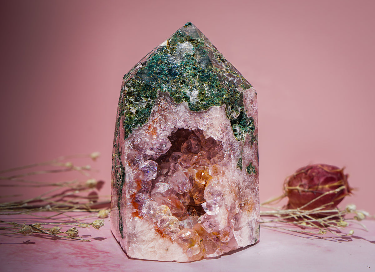 A natural pink amethyst and jasper crystal tower pointed crystal cave specimen with green and pink mineral formations, displayed on a pink surface with dried floral stems against a solid pink background.