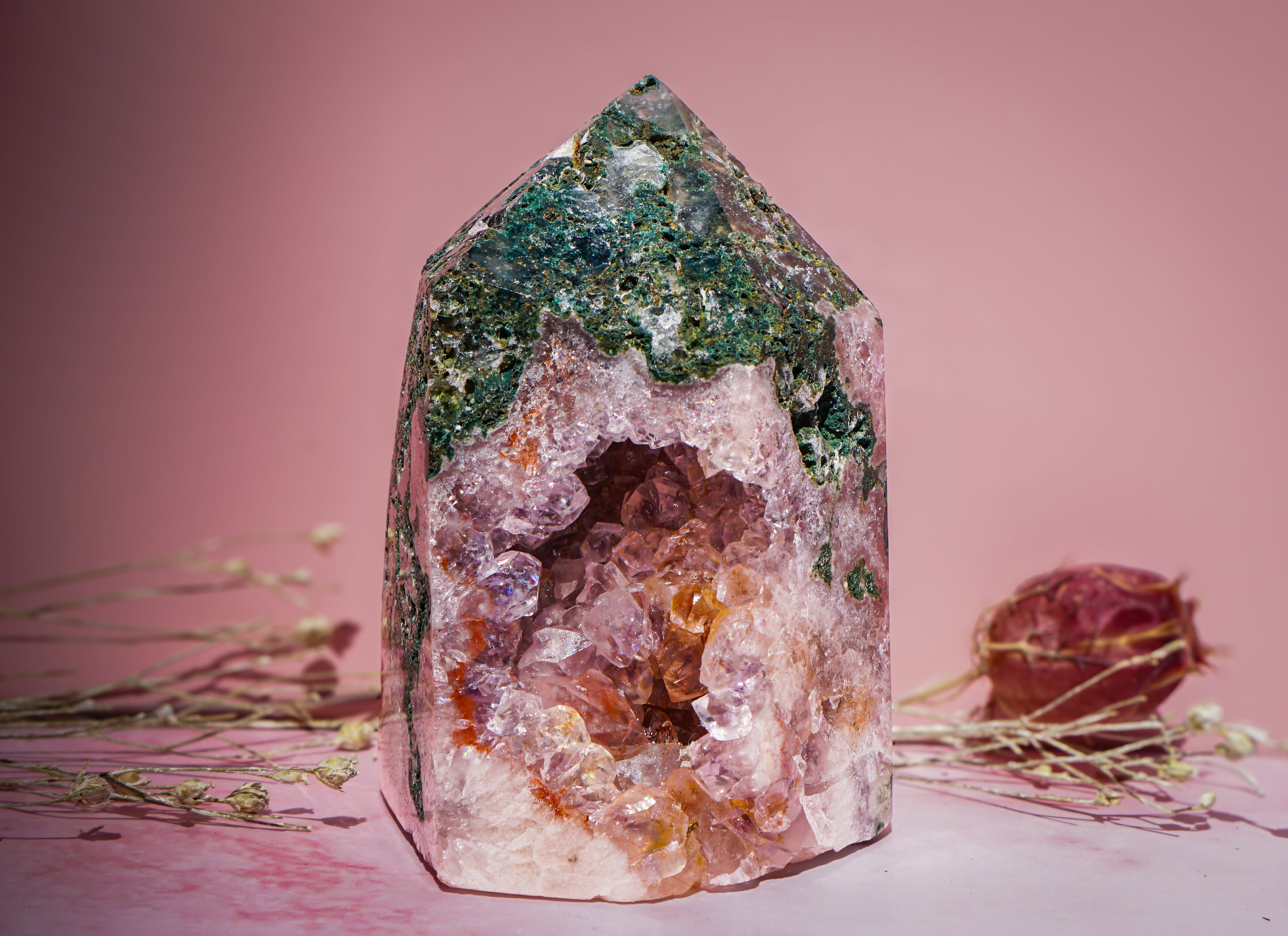 A natural pink amethyst and jasper crystal tower pointed crystal cave specimen with green and pink mineral formations, displayed on a pink surface with dried floral stems against a solid pink background.