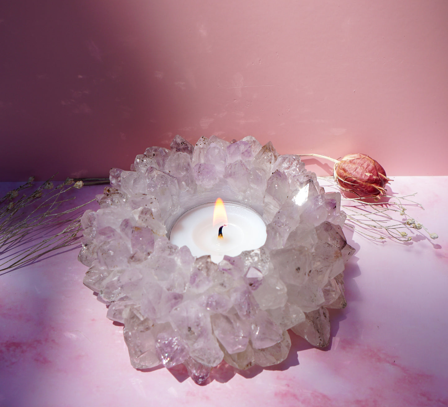 A lit white tea light candle set in a druzy brazilian amethyst crystal tea light holder, styled on a pink surface with dried botanicals against a pink backdrop.