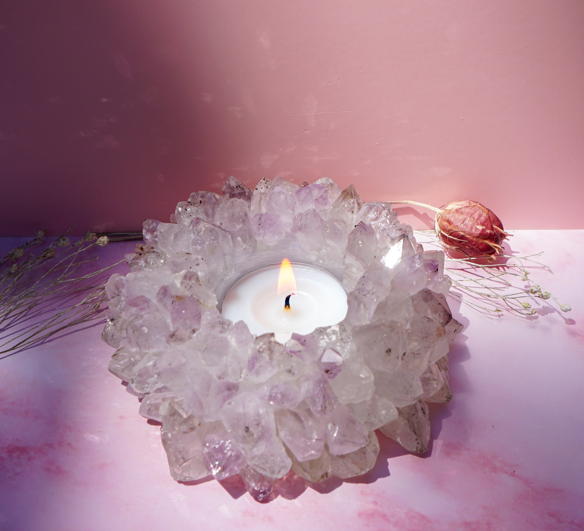 A lit white tea light candle set in a druzy brazilian amethyst crystal tea light holder, styled on a pink surface with dried botanicals against a pink backdrop.